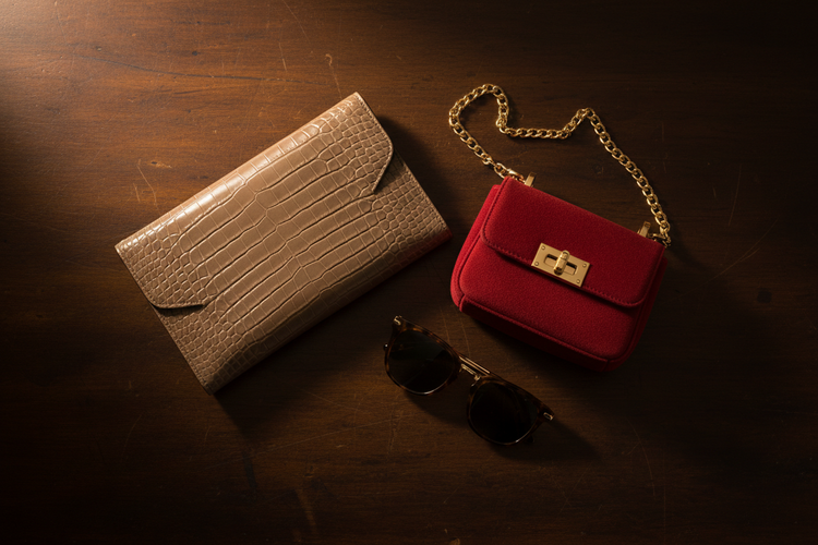 Made in Italia Kollektion – taupefarbene Leder-Clutch mit Croco-Prägung, rote Mini-Handtasche mit goldener Kette und Schildpatt-Sonnenbrille auf dunklem Holzhintergrund